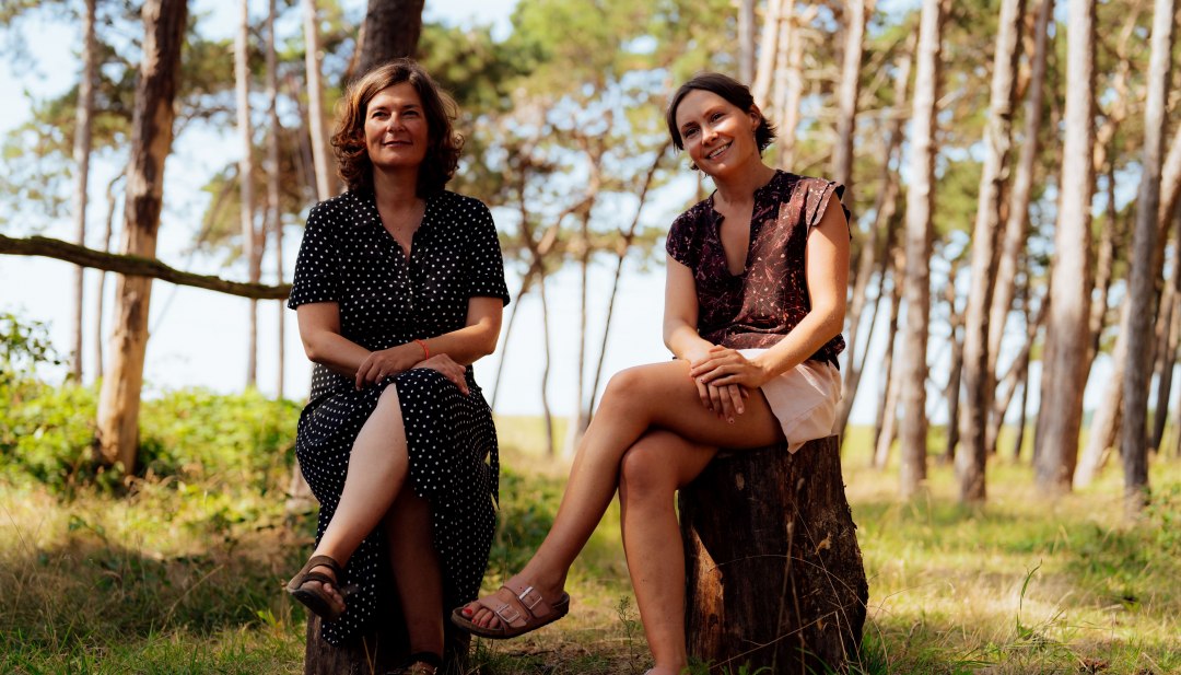 Zwei Frauen sitzen im lichten Küstenwald auf Baumstümpfen, im Hintergrund schimmern Dünen und Meer. // Anne Liedtke und Maria Decker sind in die Insel Rügen verliebt – und wollen das weitergeben. // © MV-T/Petermann Zwei Frauen sitzen im lichten Küstenwald auf Baumstümpfen, im Hintergrund schimmern Dünen und Meer.