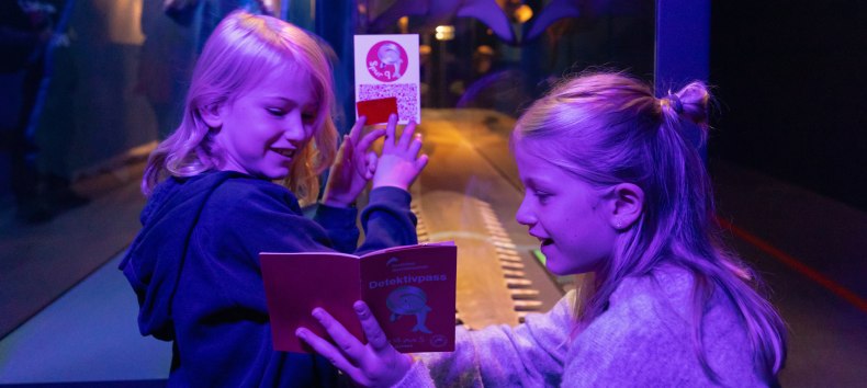 Zwei Kinder sitzen unter UV-Licht vor einer Vitrine und schauen erstaunt auf ein kleines Heft, welches eines der Kinder in den Händen hält. // © Anke Neumeister/Deutsches Meeresmuseum Zwei Kinder sitzen unter UV-Licht vor einer Vitrine und schauen erstaunt auf ein kleines Heft, welches eines der Kinder in den Händen hält. // © Anke Neumeister/Deutsches Meeresmuseum