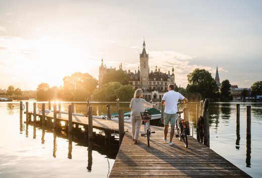 Ein Paar mit Fahrr&auml;dern steht auf einem Steg am Schweriner See und blickt im Abendlicht auf das Schweriner Schloss.