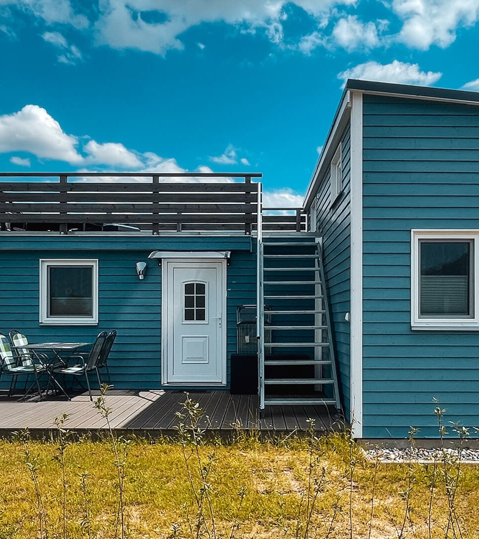 Modernes, blaues Tiny House mit Dachterrasse und gem&uuml;tlicher Holzterrasse mit Sitzbereich, umgeben von l&auml;ndlicher Landschaft unter blauem Himmel.