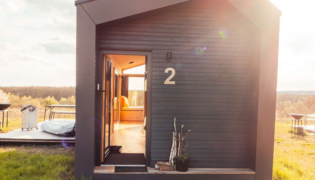 Klein, donker huisje met houten lambrisering, open ingangsdeur en zonnestralen op een groene weide.