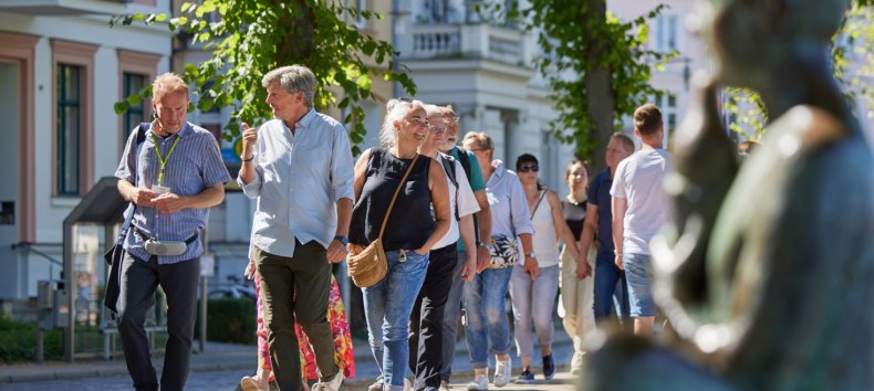 Eine Gruppe Menschen ist mit einem Stadtf&uuml;hrer auf einem gepflasterten Weg unterwegs. Rechts, unscharf im Bild ist eine Statue am Pfaffenteich zu sehen, im Hintergrund erscheinen die Villen. Blickpunkt sind der Stadtf&uuml;hrer sowie ein Paar, dass sich angeregt unterh&auml;lt. // &copy; Oliver Borchert