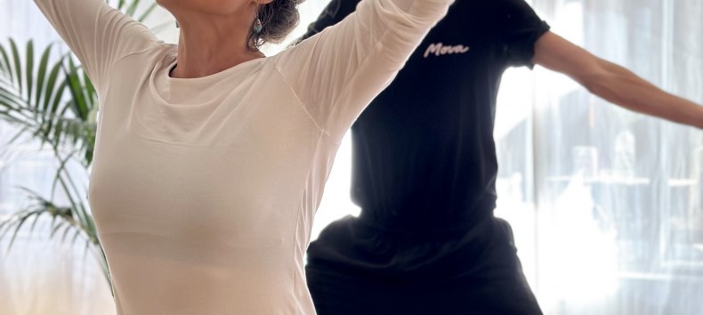 Zwei Personen praktizieren Yoga in einem hellen Studio. Im Vordergrund steht eine Frau in einer kraftvollen stehenden Yoga-Haltung mit erhobenen Armen, im Hintergrund folgt ein Mann derselben Bewegung. Weiches Tageslicht und eine ruhige Atmosph&auml;re unterstreichen Achtsamkeit, Bewegung und K&ouml;rperbewusstsein., &copy; MOVA PLACE - Yoga & Co. Studio