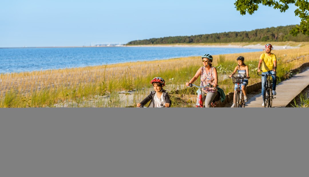 Eine vierk&ouml;pfige Familie f&auml;hrt mit dem Fahrrad auf dem Radweg am Strand entlang.
