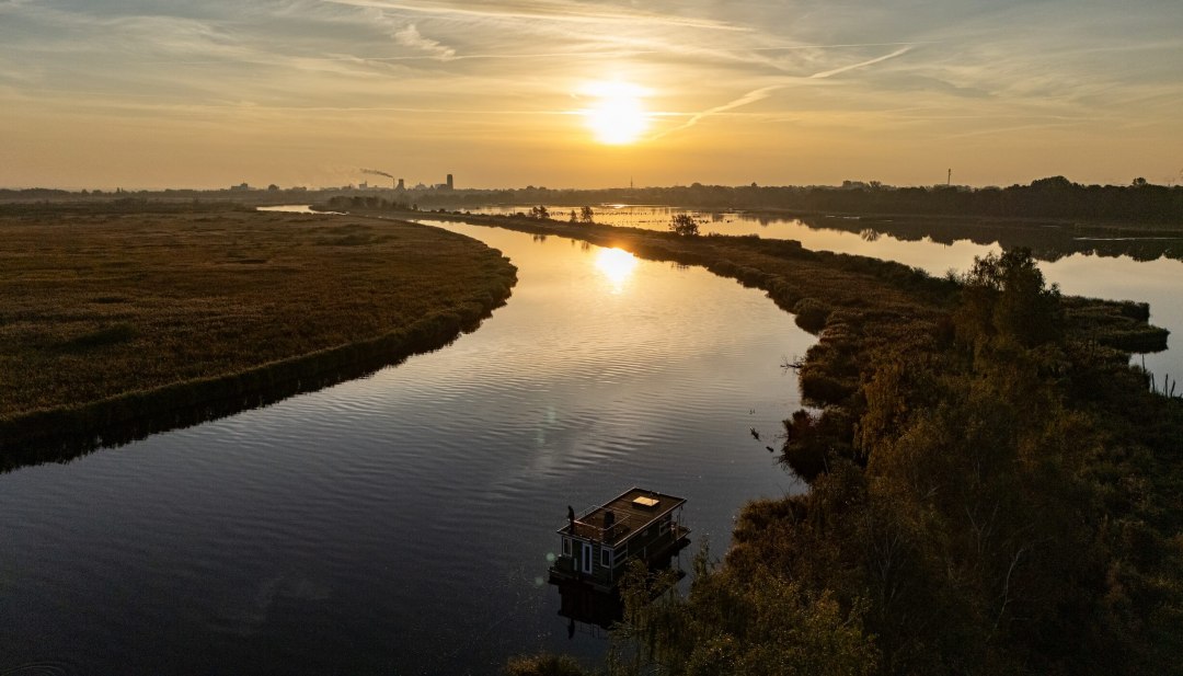 Woonboot op de Peene bij zonsondergang, omgeven door een vredig rivierlandschap en gouden licht aan de horizon.