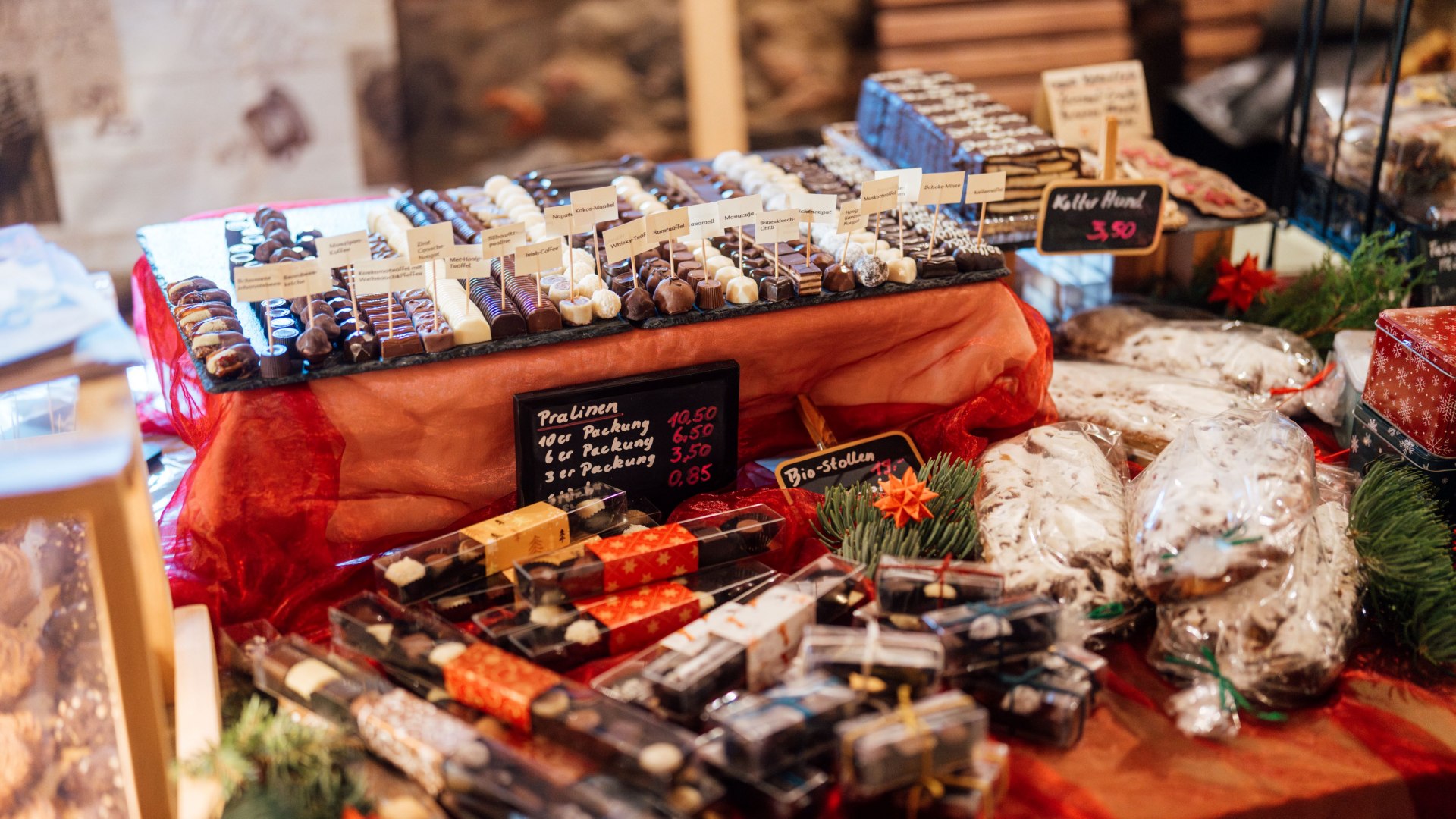 Ein festlich dekorierter Marktstand mit handgefertigten Pralinen, Schokoladen und Christstollen, umgeben von weihnachtlichem Schmuck. // Feinste Pralinen und traditioneller Christstollen – süße Verlockungen auf dem Weihnachtsmarkt laden zum Genießen ein. // © MV-T/Petermann Ein festlich dekorierter Marktstand mit handgefertigten Pralinen, Schokoladen und Christstollen, umgeben von weihnachtlichem Schmuck.