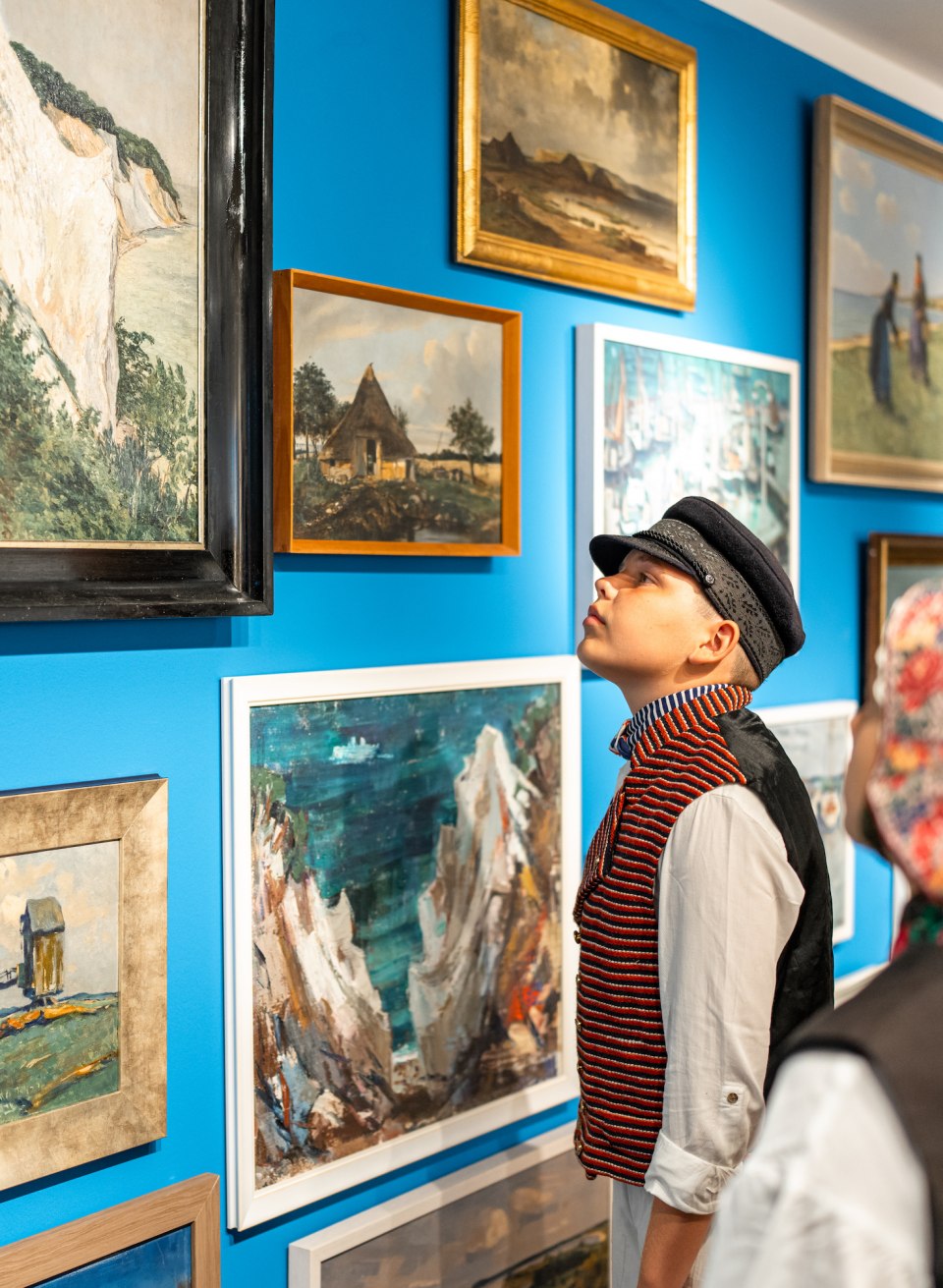 Auch Maler der Region Mönchsgut sind in der “St. Petersburger Hängung” im Göhrener Heimatmuseum vertreten, © TMV/Gross Zeichnungen und Malereien hängen an der Wand im Heimatmuseum Mönchgut - Kinder stehen in traditioneller Kleidung davor und begutachten.