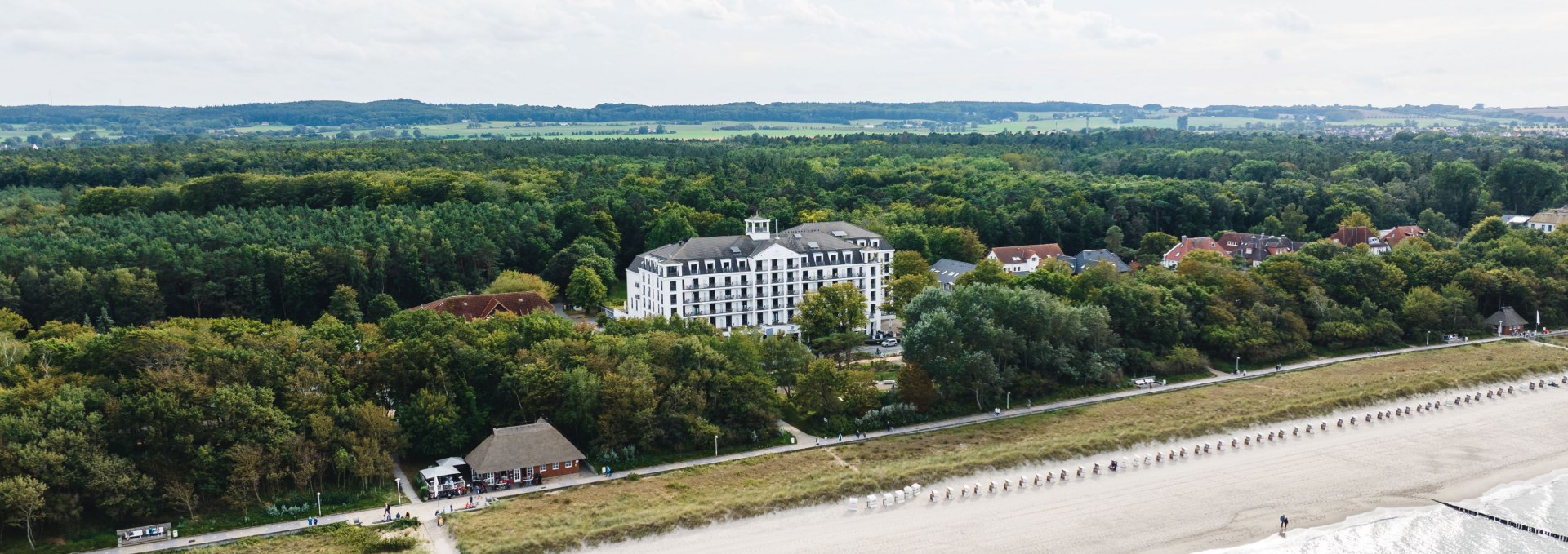 Luftfoto af stranden og et strandhotel i K&uuml;hlungsborn, omgivet af skov, med strandkurve og det rolige &Oslash;sters&oslash;vand.