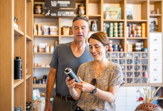 L&auml;chelndes Paar in einem Hofladen, umgeben von Regalen mit regionalen Produkten, beim Begutachten eines Artikels.
