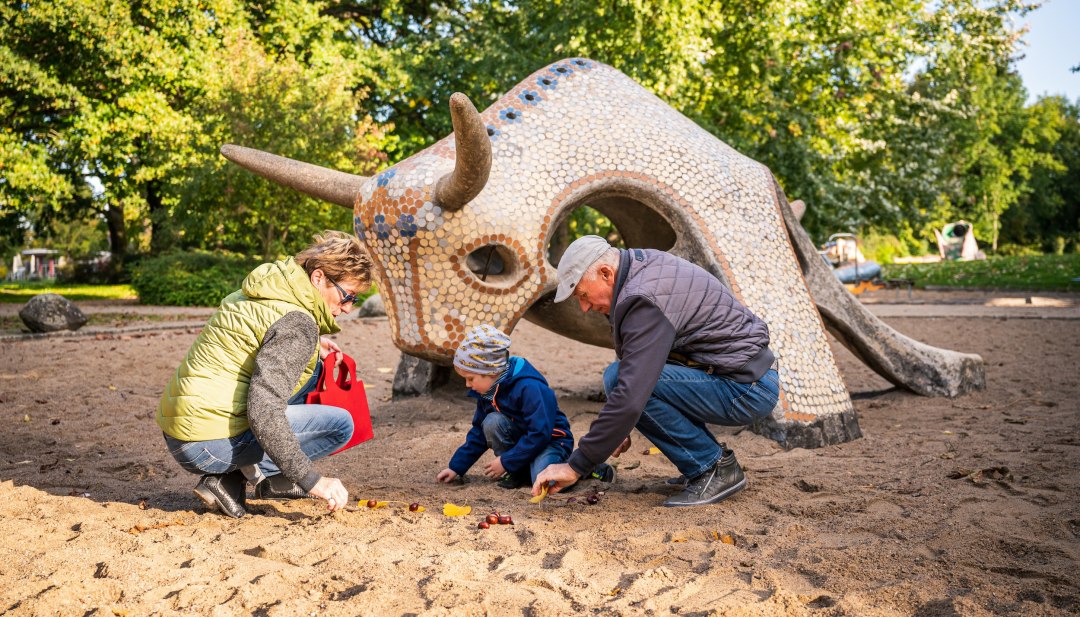 Een vrouw en een man zitten met een kind in het zand voor een betegelde sculptuur van een stier en zoeken kastanjes. // Kastanjes in het zand, bladeren in de hand: op de speeltuin aan het Tollensemeer in Neubrandenburg genieten gezinnen samen van de herfst. // © MV-T/Tiemann Een vrouw en een man zitten met een kind in het zand voor een betegelde sculptuur van een stier en zoeken kastanjes.