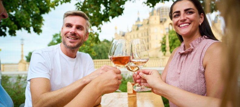 G&auml;ste sto&szlig;en beim Winzerfest Schwerin auf dem Alten Garten mit einem Glas Wein an. Im Hintergrund erhebt sich das Schweriner Schloss und gibt dem Weinfest seine besondere Kulisse. // &copy; LHSN/Celien Graubaum