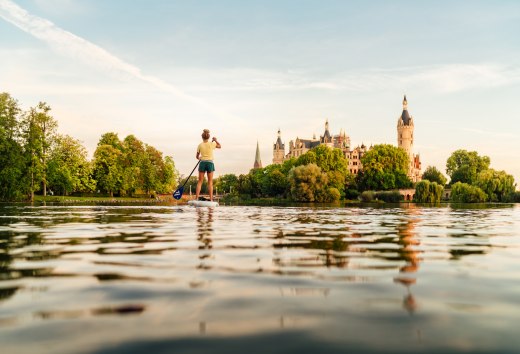 Beim Gleiten über den Schweriner See entsteht ein stiller, kraftvoller Augenblick, in dem Natur und Schlosskulisse miteinander verschmelzen., © MV-T/Petermann Eine Person paddelt bei warmem Abendlicht auf einem See und blickt auf ein historisches Schloss, umgeben von üppigem Grün.
