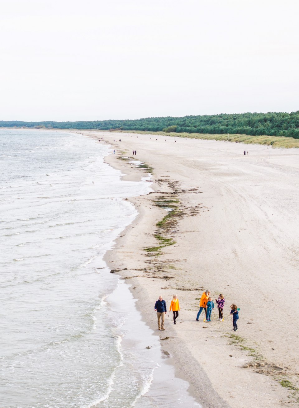 Luftaufnahme eines langen, sandigen Strandes, an dessen Ufer eine Gruppe von Menschen spaziert, umgeben von Meer und gr&uuml;nen D&uuml;nen.