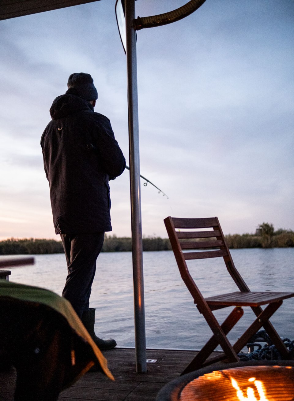 Ein Angler genießt den ruhigen Abend auf einem Hausbootdeck an der Peene, während ein wärmendes Feuer flackert., © TMV/Scholz-Witzel Person angelt vom Deck eines Hausboots auf der Peene bei Sonnenuntergang, mit Feuerstelle im Vordergrund.