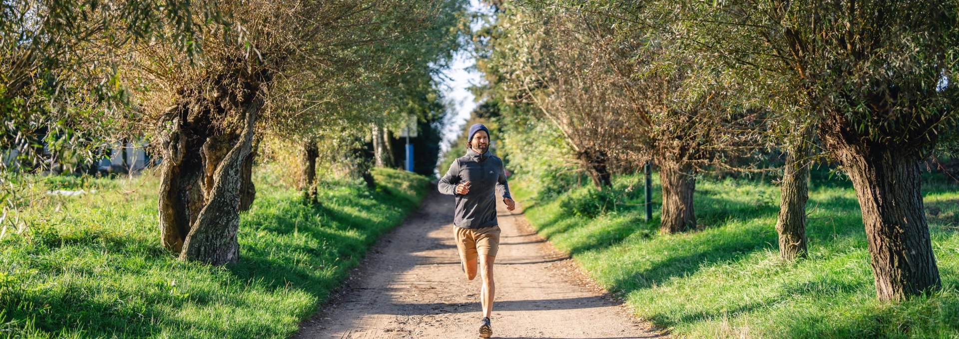  Ein Mann joggt auf einem von B&auml;umen ges&auml;umten Weg auf der Insel Poel, umgeben von gr&uuml;ner Landschaft und sonnigem Wetter.