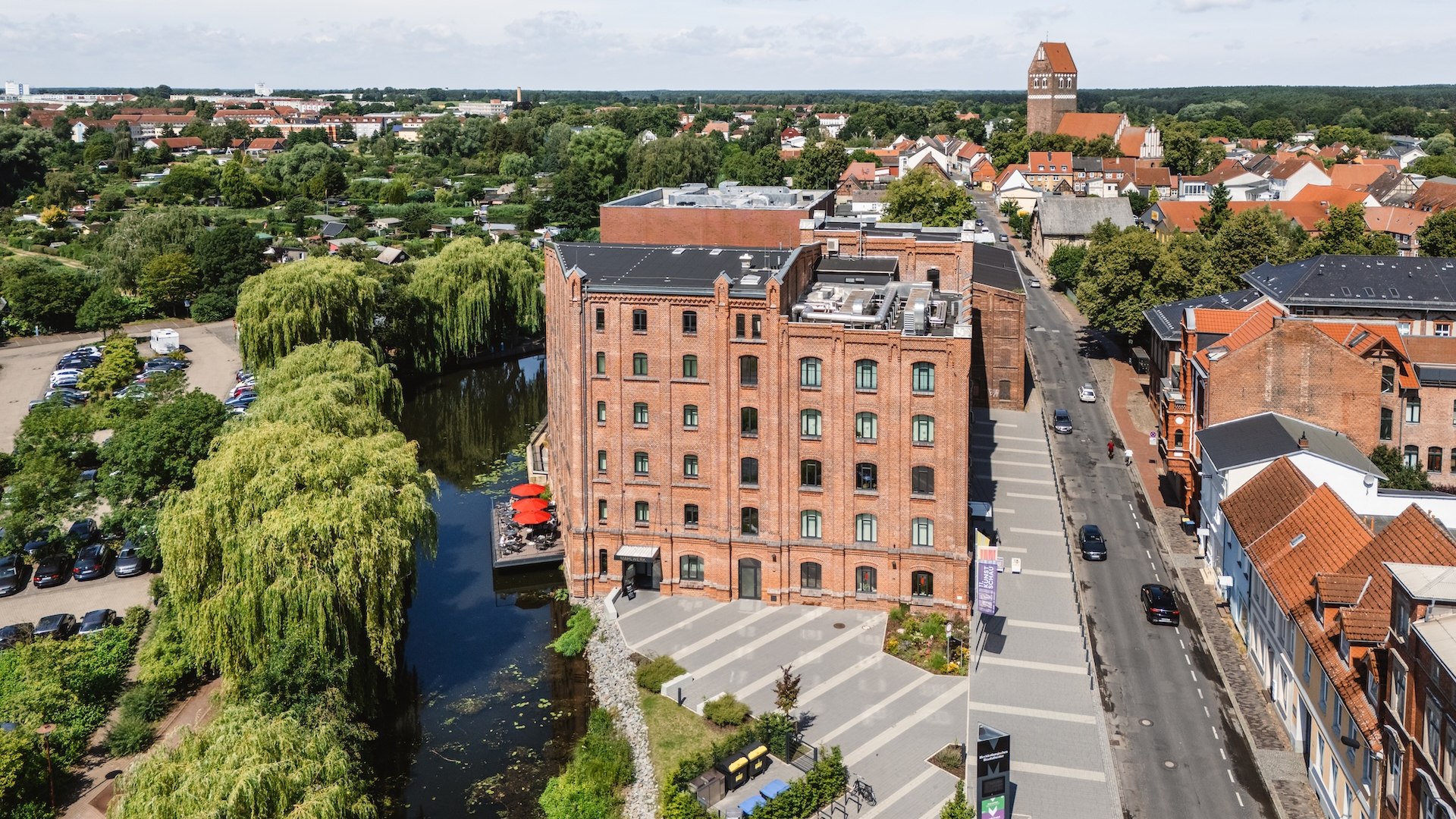 Eine Luftaufnahme zeigt die Kulturm&uuml;hle in Parchim mit ihrer historischen Fassade und einer gem&uuml;tlichen Restaurantterrasse am Wasser, umgeben von viel Gr&uuml;n.