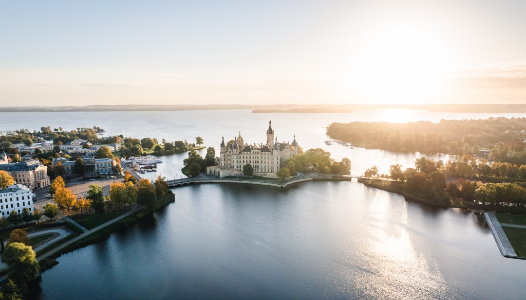 Schweriner Schloss auf Insel im See bei Sonnenaufgang, umgeben von B&auml;umen und Wasserfl&auml;chen.