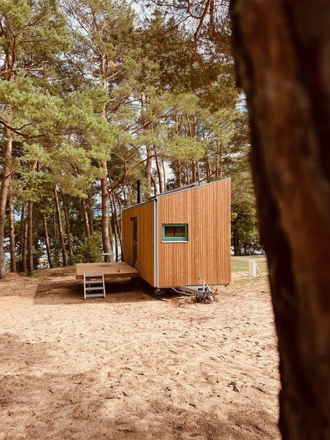 Freistehendes Tiny House mit Holzverkleidung auf sandigem Boden, umgeben von hohen Kiefern in einer idyllischen Waldlandschaft. // Nachhaltige Tiny Houses in der Natur // © Ahoi Camp Canow Freistehendes Tiny House mit Holzverkleidung auf sandigem Boden, umgeben von hohen Kiefern in einer idyllischen Waldlandschaft.