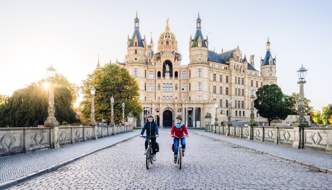 Twee fietsers steken een brug over voor het kasteel van Schwerin, verlicht door de gouden ochtendzon.