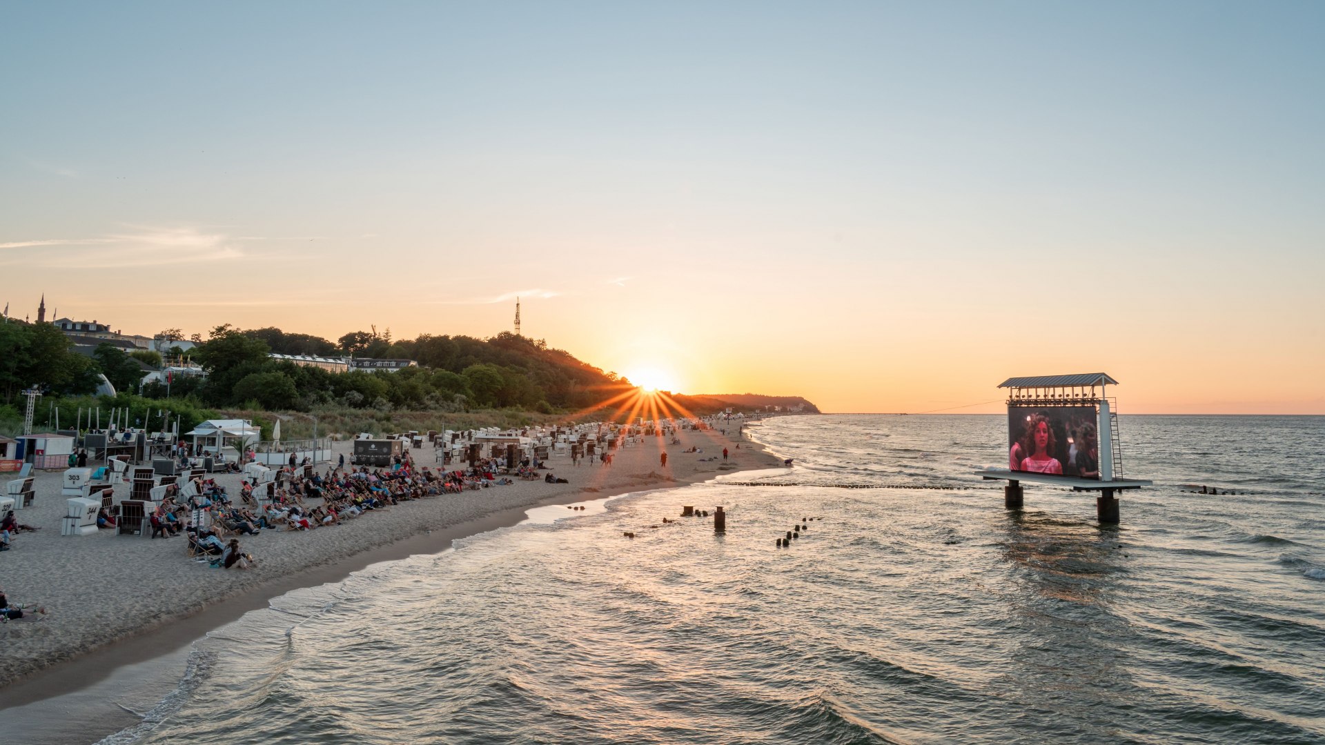 Strandkino-Event auf Usedom mit LED-Leinwand im Meer und Sonnenuntergang hinter Zuschauern am Ostseestrand.