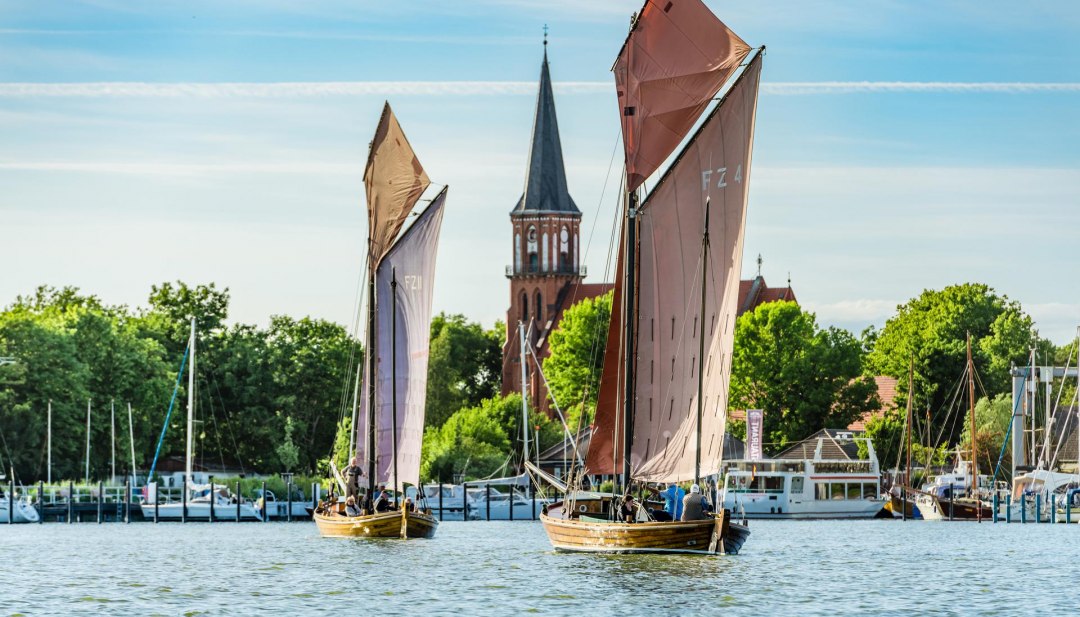 Zwei Zeesenboote segeln auf dem Bodden vor dem Hafen Wustrow mit Blick auf die Dorfkirche. // © MV-T/Tiemann Zwei Zeesenboote segeln auf dem Bodden vor dem Hafen Wustrow mit Blick auf die Dorfkirche.