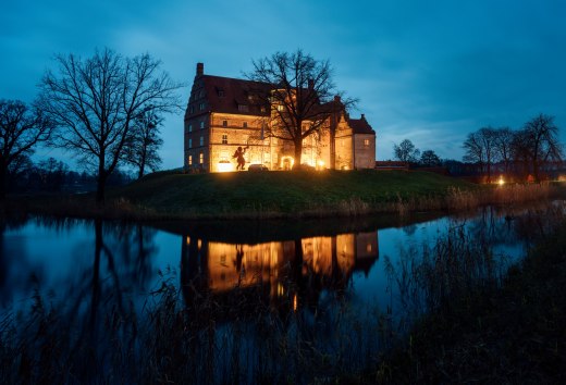 Weihnachtliche Idylle am Schloss Ulrichshusen – festliche Beleuchtung spiegelt sich im Wassergraben und sorgt für eine märchenhafte Abendstimmung., © TMV/Petermann Stimmungsvoll beleuchtetes Schloss Ulrichshusen zur Weihnachtszeit, umgeben von Bäumen und Wassergraben mit Spiegelung im Abendlicht.