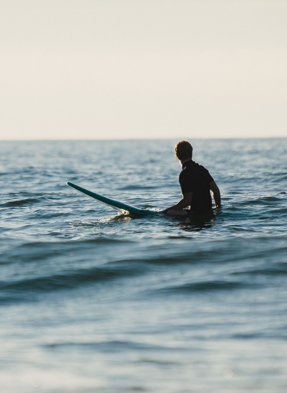 Eine Person sitzt mit Surfboard in den sanften Ostseewellen vor der K&uuml;ste von Warnem&uuml;nde und blickt &uuml;ber das ruhige Wasser.