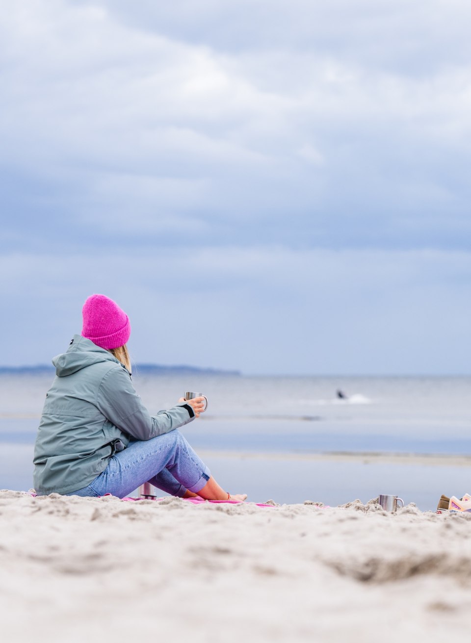  Een vrouw met een roze pet zit op het strand van Timmendorf, kijkt uit over zee en houdt een kopje in haar hand.