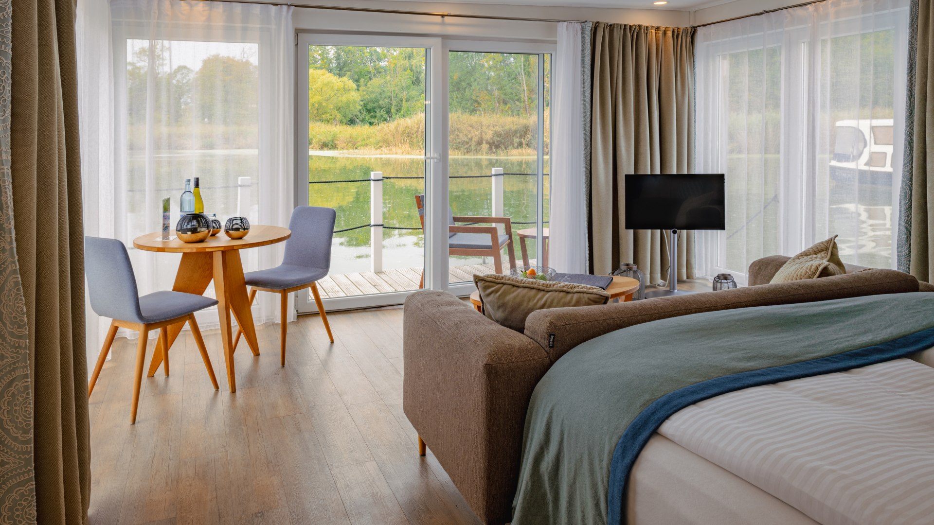 Elegantes Interieur und ungetrübter Ausblick: Die schwimmenden Suiten im Naturhafen Krummin, © TMV/Tiemann Die schwimmenden Suiten im Naturhafen Krummin von Innen mit Couch, TV und Blick auf Terrasse mit Sitzgelegenheiten