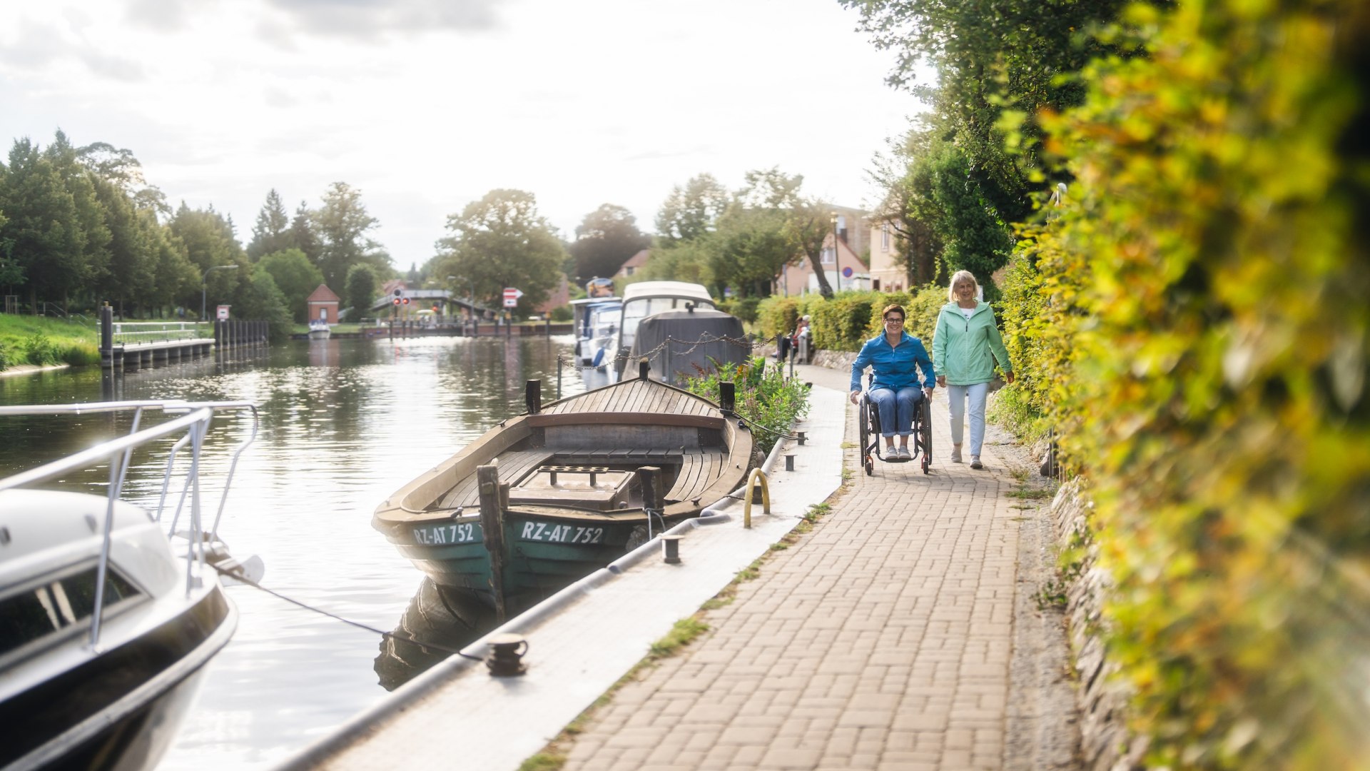 Eine Rollstuhlfahrerin unterwegs mit Ihrer Freundin am Kanal in Plau am See. An der Kante liegen Boote.