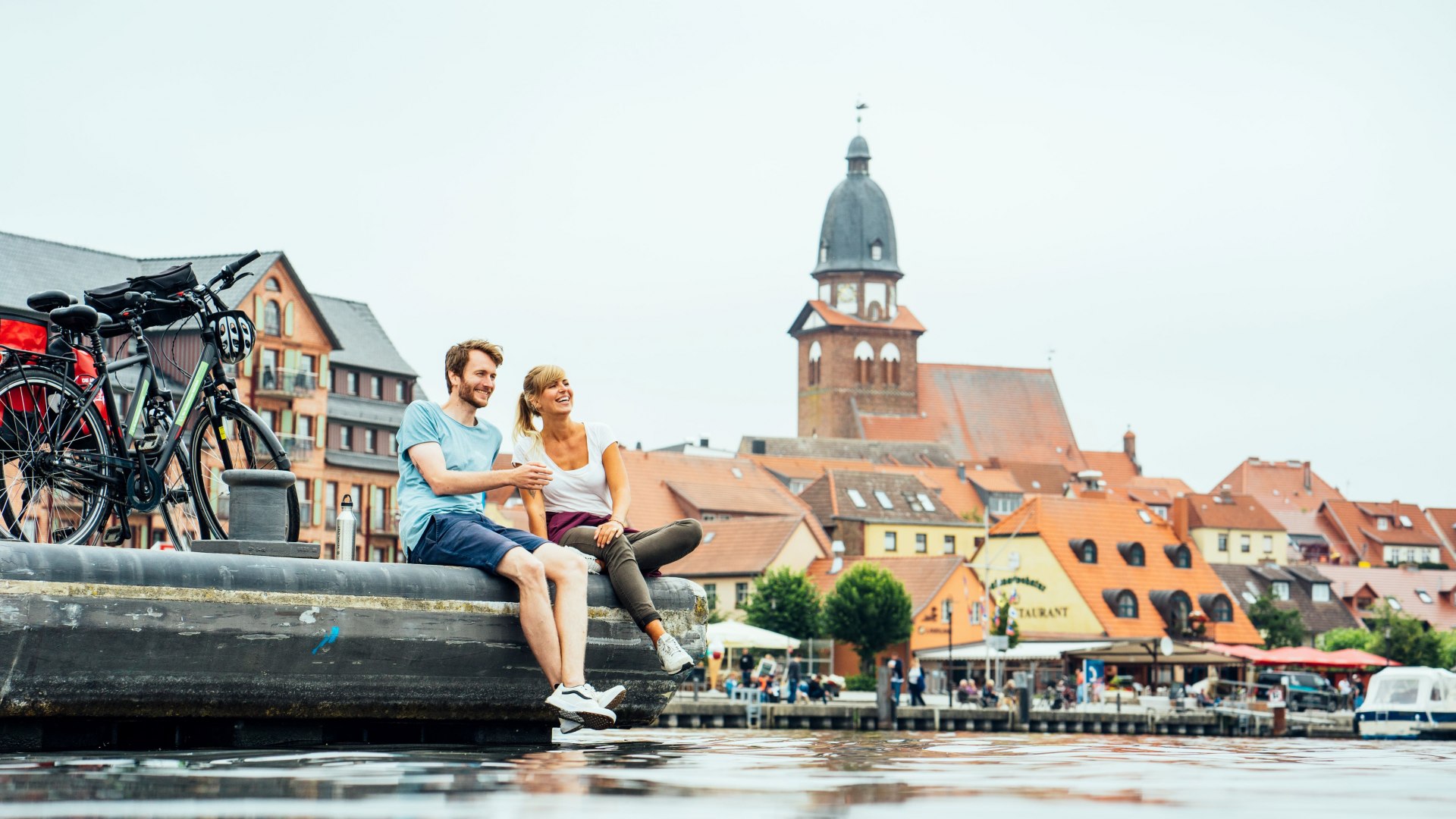 Ein junges Paar sitzt am Wasser im Hafen von Waren (Müritz), Fahrräder daneben, im Hintergrund die Altstadt mit Kirchturm.