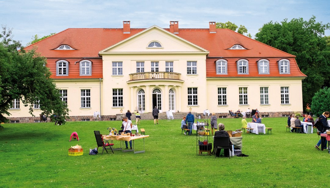 Menschen sitzen bei einer Mittsommerveranstaltung auf der Wiese vor dem Gutshaus Gevezin, w&auml;hrend das historische Geb&auml;ude hell im Tageslicht strahlt.