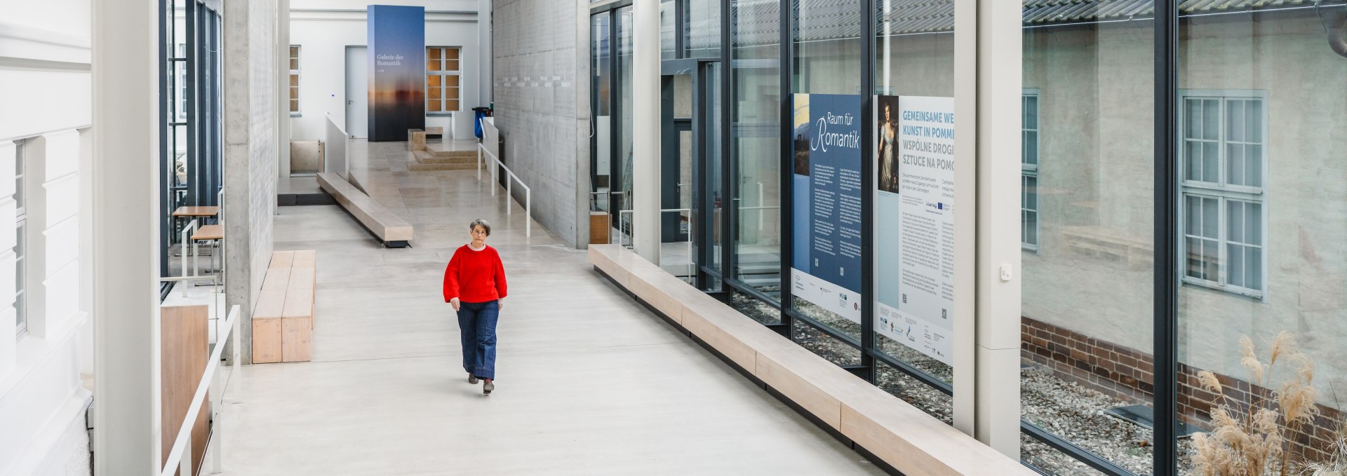 Lichte museumgang met glazen gevel in het Pommers Staatsmuseum Greifswald, bezoeker in rode trui.