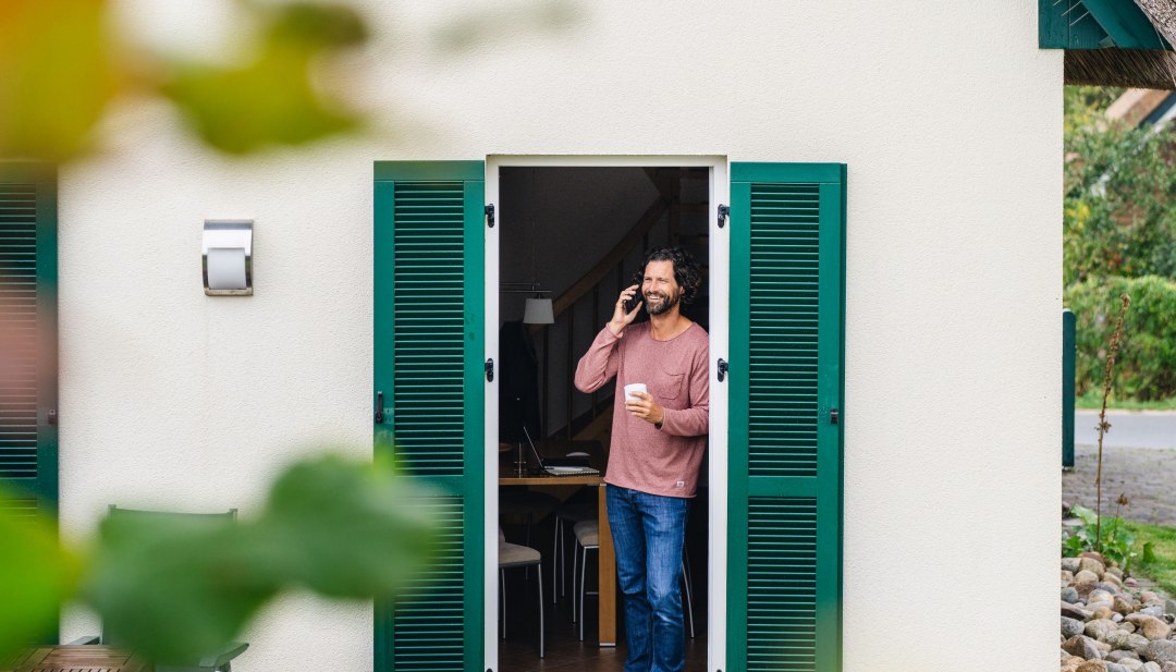 Mann telefoniert mit Kaffeebecher in der Hand an der T&uuml;r eines Ferienhauses auf der Insel Poel w&auml;hrend seiner Workation.