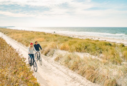 Twee mensen fietsen over een zandduinpad aan de kust van Hiddensee, terwijl de golven op het strand slaan en het gras wiegt in de wind.