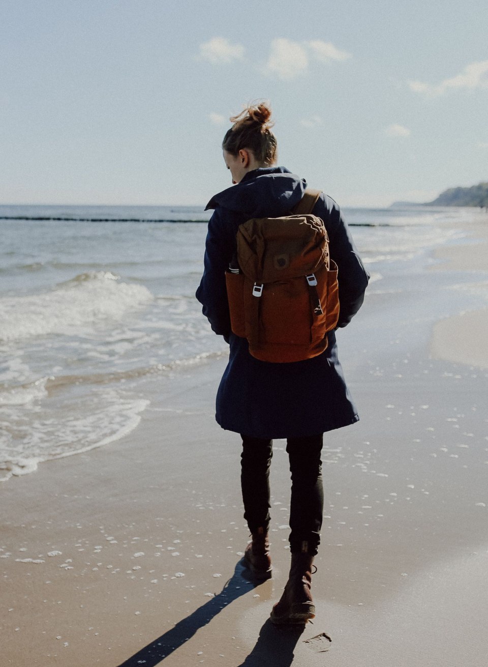 Eine Spazierg&auml;ngerin wandert entlang der K&uuml;ste der Insel Usedom, w&auml;hrend die sanften Wellen den Strand umsp&uuml;len. Mit einem Rucksack ausgestattet, begibt sie sich auf die Suche nach verborgenen Sch&auml;tzen wie Bernsteinen, die hier oft an den Ufern zu finden sind.