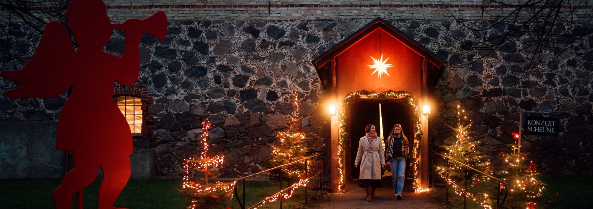 Zwei Frauen gehen aus der weihnachtlich beleuchteten Konzertscheune von Schloss Ulrichshusen, flankiert von Lichterbäumen und einer Engelsfigur. // Stimmungsvolle Adventszeit – Besucherinnen verlassen die festlich geschmückte Konzertscheune von Schloss Ulrichshusen nach einer Weihnachtsveranstaltung. // © MV-T/Petermann Zwei Frauen gehen aus der weihnachtlich beleuchteten Konzertscheune von Schloss Ulrichshusen, flankiert von Lichterbäumen und einer Engelsfigur.