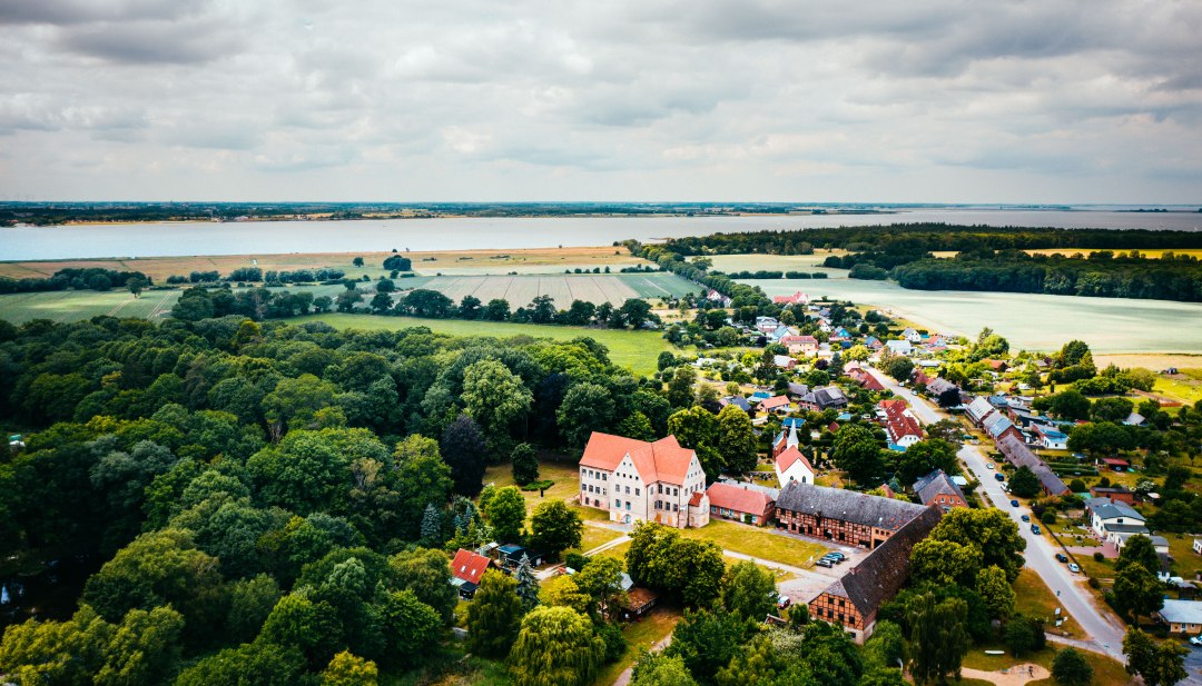 Das Schloss Ludwigsburg mit rotem Dach ragt aus dem grünen Dorf, dahinter reicht der Blick über Felder bis zum Wasser. // Zwischen alten Bäumen und Feldern liegt das Schloss Ludwigsburg, der Blick reicht bis zum Wasser am Horizont. Hier lässt sich der Tag entschleunigen und die weite Landschaft erkunden. // © MV-T/Friedrich Das Schloss Ludwigsburg mit rotem Dach ragt aus dem grünen Dorf, dahinter reicht der Blick über Felder bis zum Wasser.