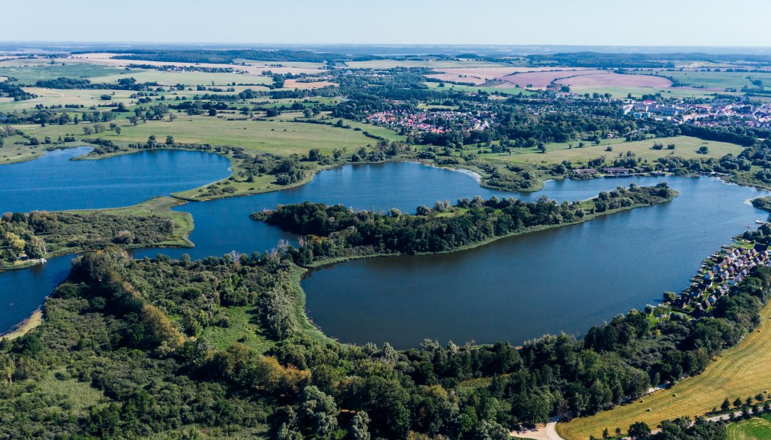Luftaufnahme vom Teterower See mit Burgwallinsel, grünen Ufern, Feldern und Teterow im Hintergrund. // Weite Wasserflächen und grüne Uferlinien rahmen die Burgwallinsel im Teterower See – Raum zum Durchatmen und Erkunden in Mecklenburg-Vorpommern. // © MV-T/Gänsicke Luftaufnahme vom Teterower See mit Burgwallinsel, grünen Ufern, Feldern und Teterow im Hintergrund.