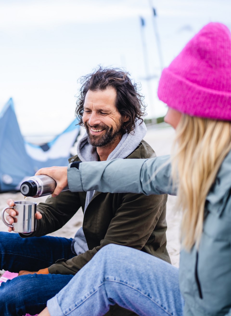 Een koppel zit op het strand in Timmendorf, glimlacht en geniet van een warme kop koffie met een vlieger op de achtergrond.