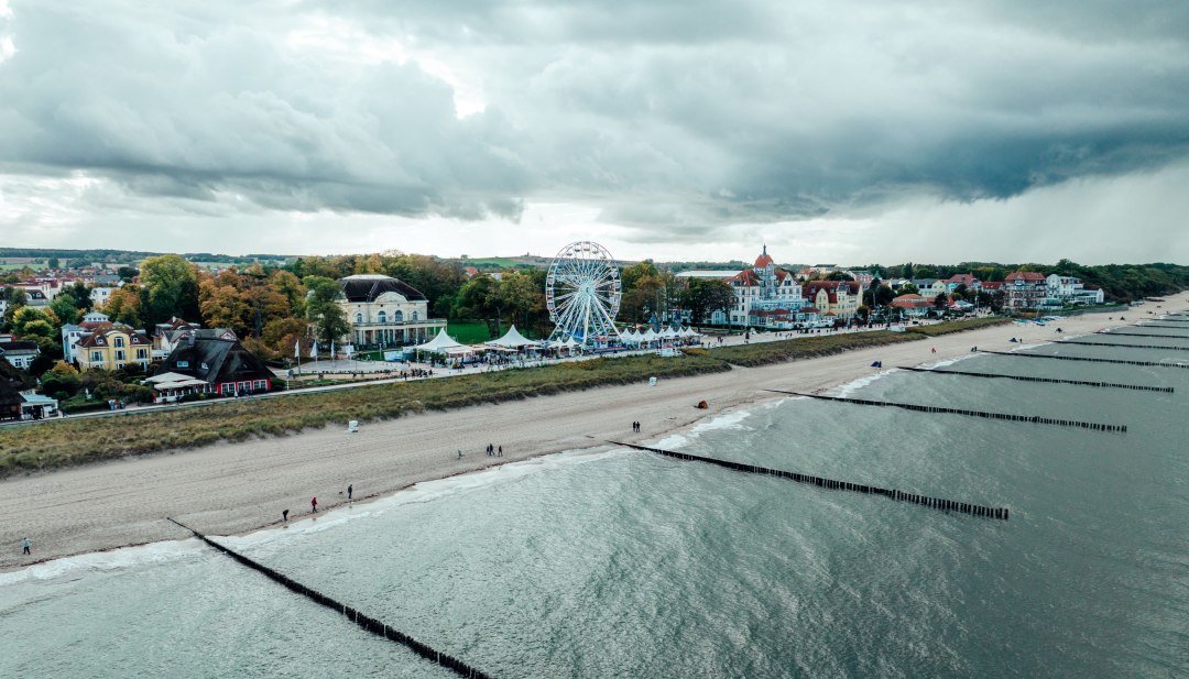 Luftaufnahme der Strandpromenade von K&uuml;hlungsborn an der Ostsee mit Sandstrand, Buhnen, Riesenrad und historischen Geb&auml;uden im Hintergrund.