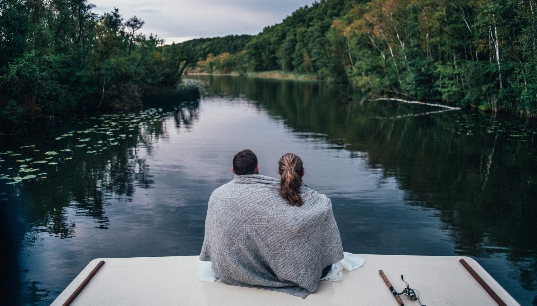 Ein Paar sitzt auf einem Hausboot, eingeh&uuml;llt in eine Decke, und blickt auf einen ruhigen, bewaldeten Fluss in der Mecklenburgischen Seenplatte.