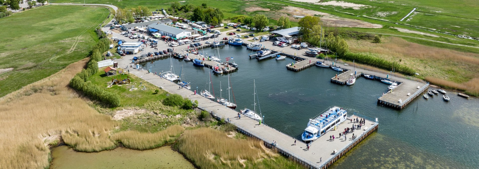 Luftaufnahme des Hafens in Thiessow mit Liegepl&auml;tzen f&uuml;r Boote und Schiffe, gr&uuml;nen Wiesen im Hintergrund und der Ostsee unter klarem Himmel.