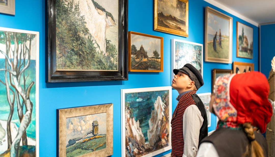 Zeichnungen und Malereien hängen an der Wand im Heimatmuseum Mönchgut - Kinder stehen in traditioneller Kleidung davor und begutachten. // Auch Maler der Region Mönchsgut sind in der “St. Petersburger Hängung” im Göhrener Heimatmuseum vertreten // © MV-T/Gross Zeichnungen und Malereien hängen an der Wand im Heimatmuseum Mönchgut - Kinder stehen in traditioneller Kleidung davor und begutachten.