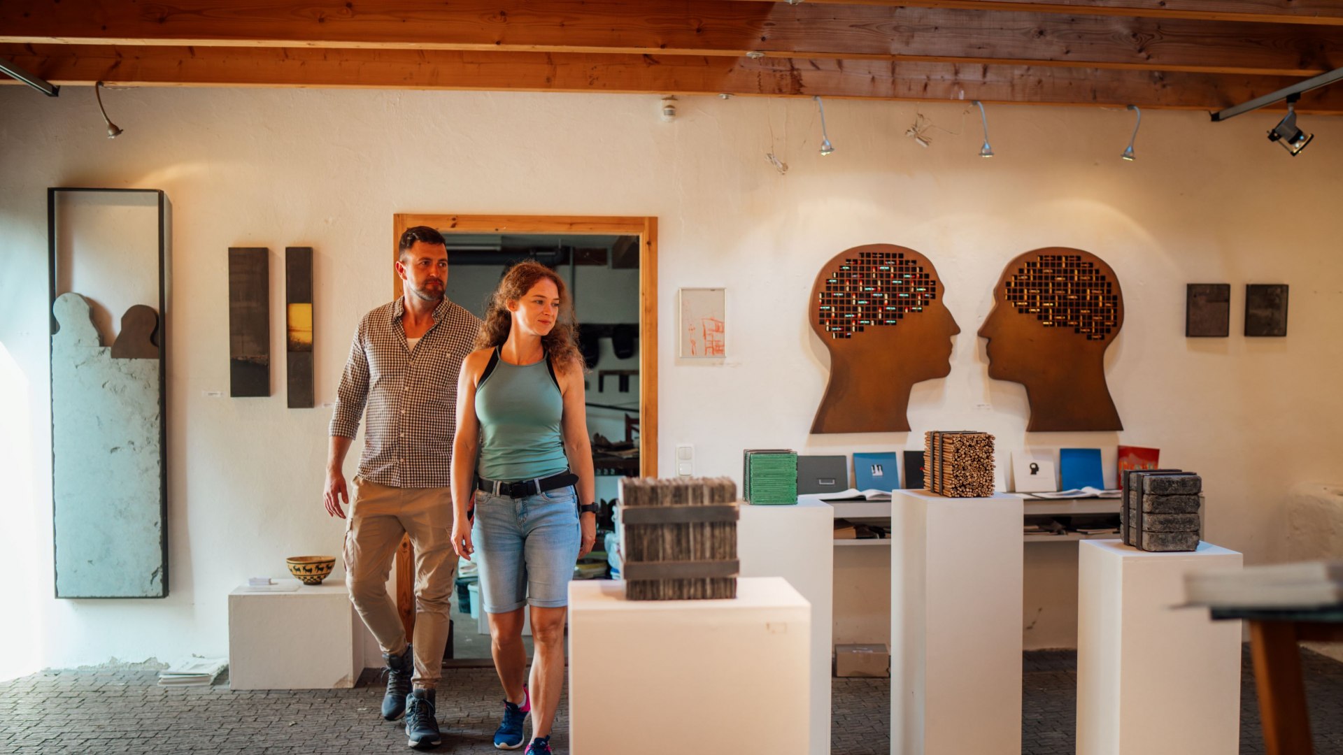 Ein Paar schlendert durch eine Kunstausstellung in der Alten Wassermühle in Wreechen. Die Ausstellung zeigt Skulpturen und Kunstwerke, darunter zwei Silhouetten menschlicher Köpfe mit detailreicher Gestaltung. Die warme Beleuchtung und die rustikale Atmosphäre schaffen eine einladende Stimmung. // Kunst entdecken in der Alten Wassermühle in Wreechen auf Rügen – moderne Skulpturen und außergewöhnliche Installationen laden zum Staunen ein. // © MV-T/Petermann Ein Paar schlendert durch eine Kunstausstellung in der Alten Wassermühle in Wreechen. Die Ausstellung zeigt Skulpturen und Kunstwerke, darunter zwei Silhouetten menschlicher Köpfe mit detailreicher Gestaltung. Die warme Beleuchtung und die rustikale Atmosphäre schaffen eine einladende Stimmung.