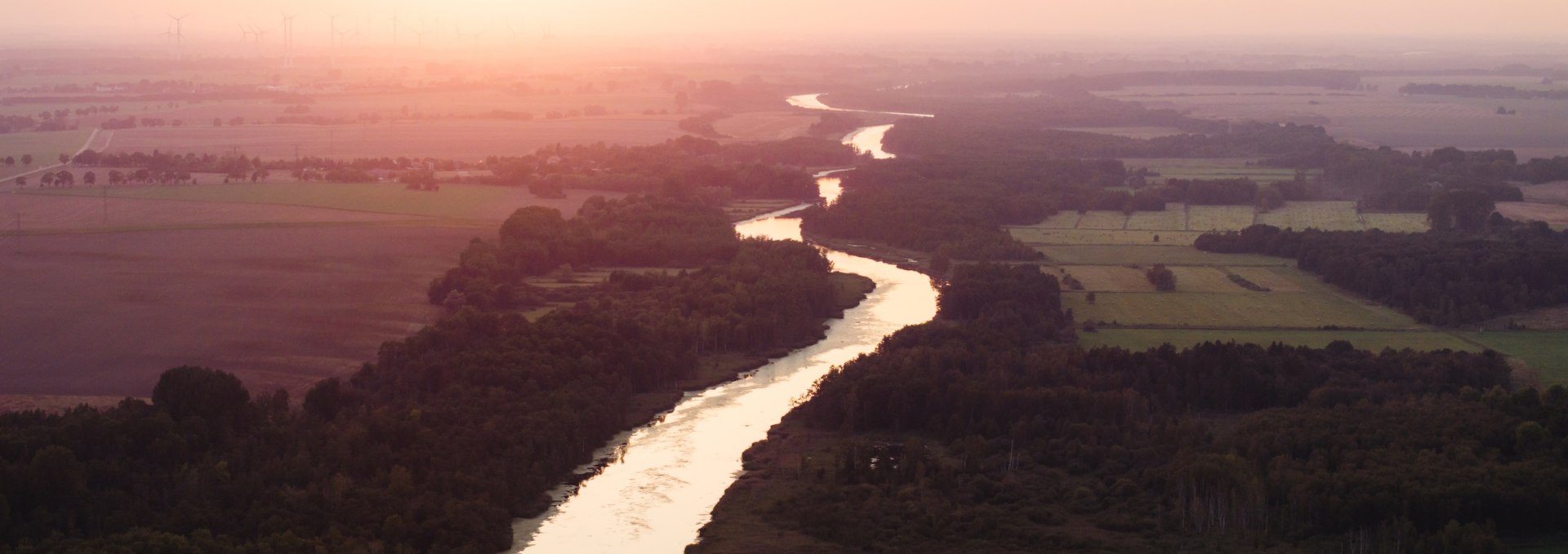 Luftaufnahme der Peene bei Sonnenuntergang, eingebettet in Felder und W&auml;lder, mit einem orangefarbenen Himmel im Hintergrund.