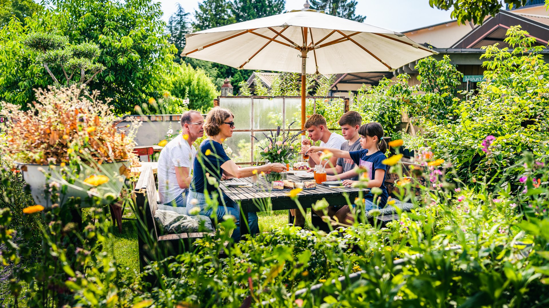 Eine Familie macht Kaffeepause im Garten des Cafés, der mit vielen Pflanzen bewachsen ist.