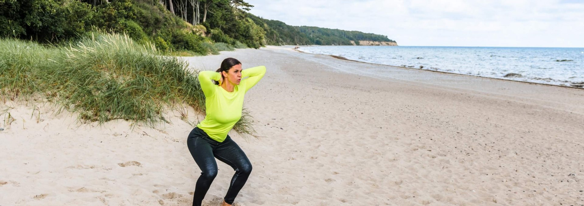 Een vrouw doet fitnessoefeningen op het strand: Squats met haar armen gekruist in haar nek.