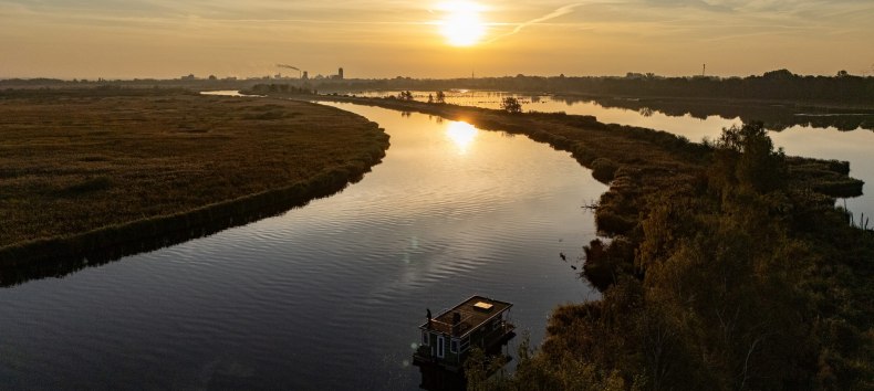 Sonnenuntergang über der Peene, während ein Hausboot friedlich auf dem Fluss treibt. Die Landschaft erstreckt sich weit in die Ferne, wo sich Himmel und Wasser treffen., © TMV Hausboot auf der Peene bei Sonnenuntergang, umgeben von ruhiger Flusslandschaft und goldenem Licht am Horizont.