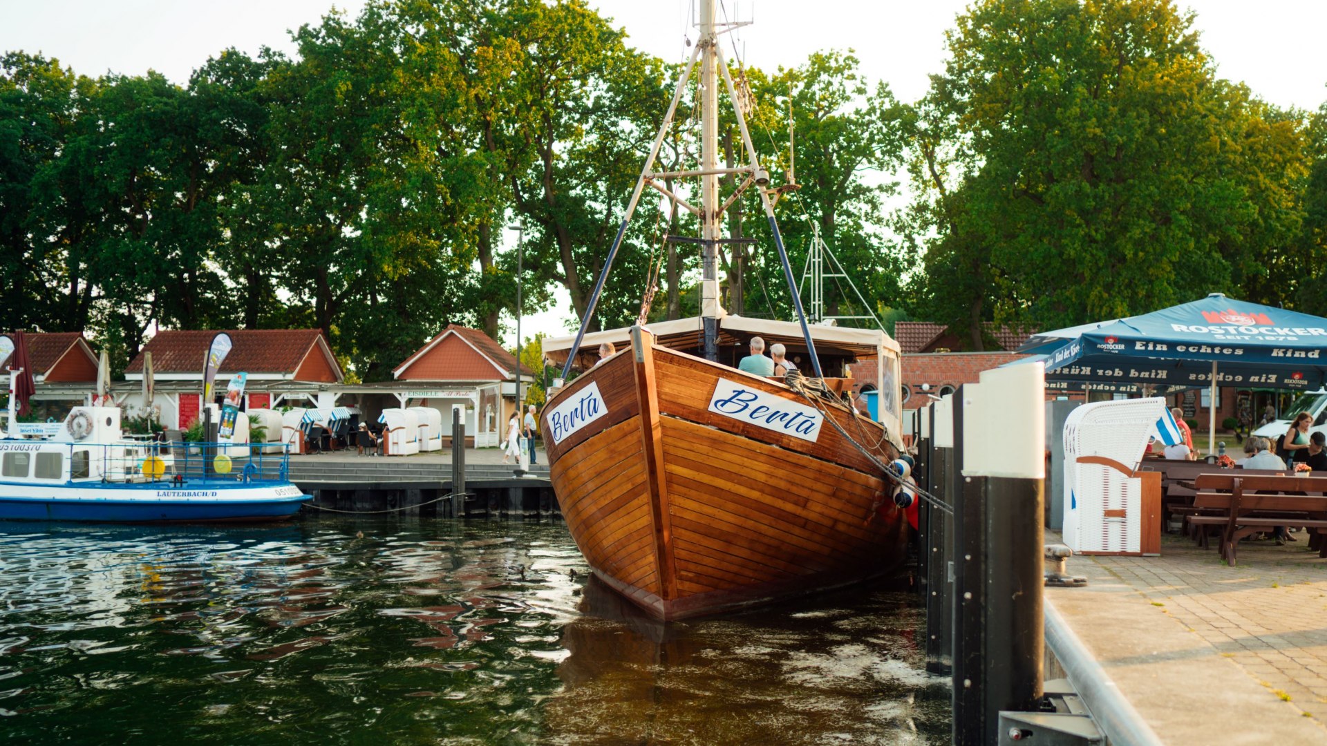 Ein h&ouml;lzerner Kutter mit dem Namen &bdquo;Berta&ldquo; liegt im Hafen von Lauterbach auf R&uuml;gen. Im Hintergrund sind kleine rote Geb&auml;ude, Strandk&ouml;rbe und Sitzbereiche unter Sonnenschirmen sichtbar. Umgeben von ruhigem Wasser und gr&uuml;nen B&auml;umen bietet die Szenerie eine idyllische Hafenatmosph&auml;re.